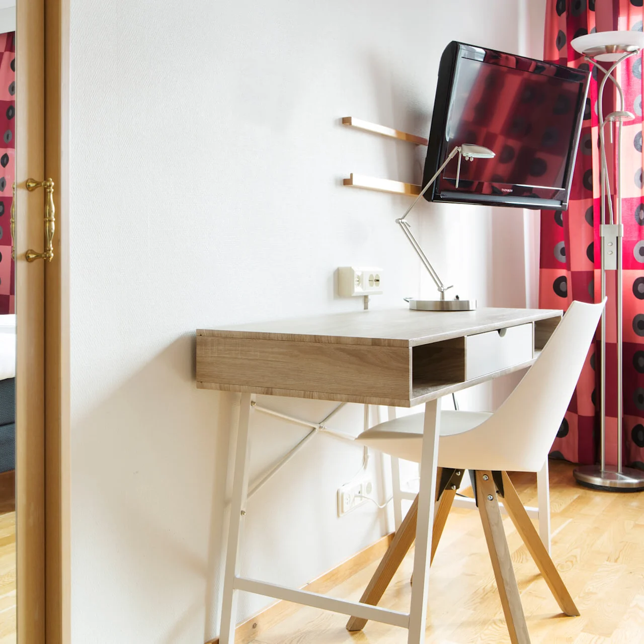 A desk with a chair stands near a wall, facing a mounted TV. Nearby, a bed with white linens is partially visible. Red curtains decorate the scene. 