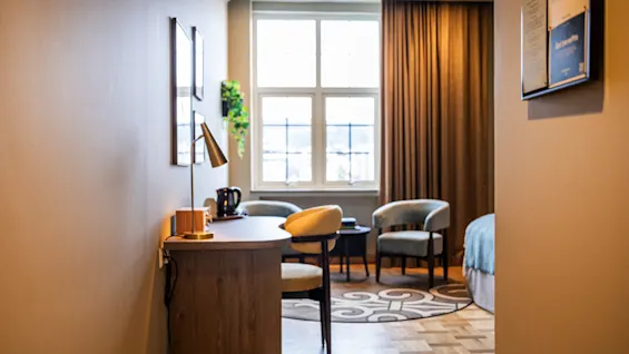 Ett mysigt hotellrum har två stolar och ett litet bord vid ett stort fönster. Ett skrivbord med stol och lampa står i närheten, och en säng är delvis synlig, med ett strukturerat parkettgolv under.