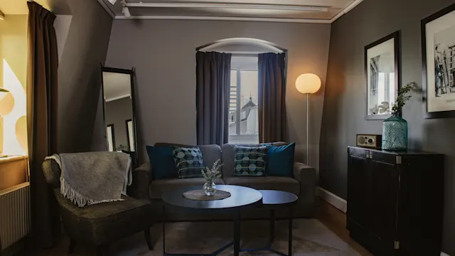 A cozy living room features a grey sofa with blue cushions and a side chair, complemented by a round coffee table. A floor lamp and framed pictures enhance the warm, inviting ambiance.