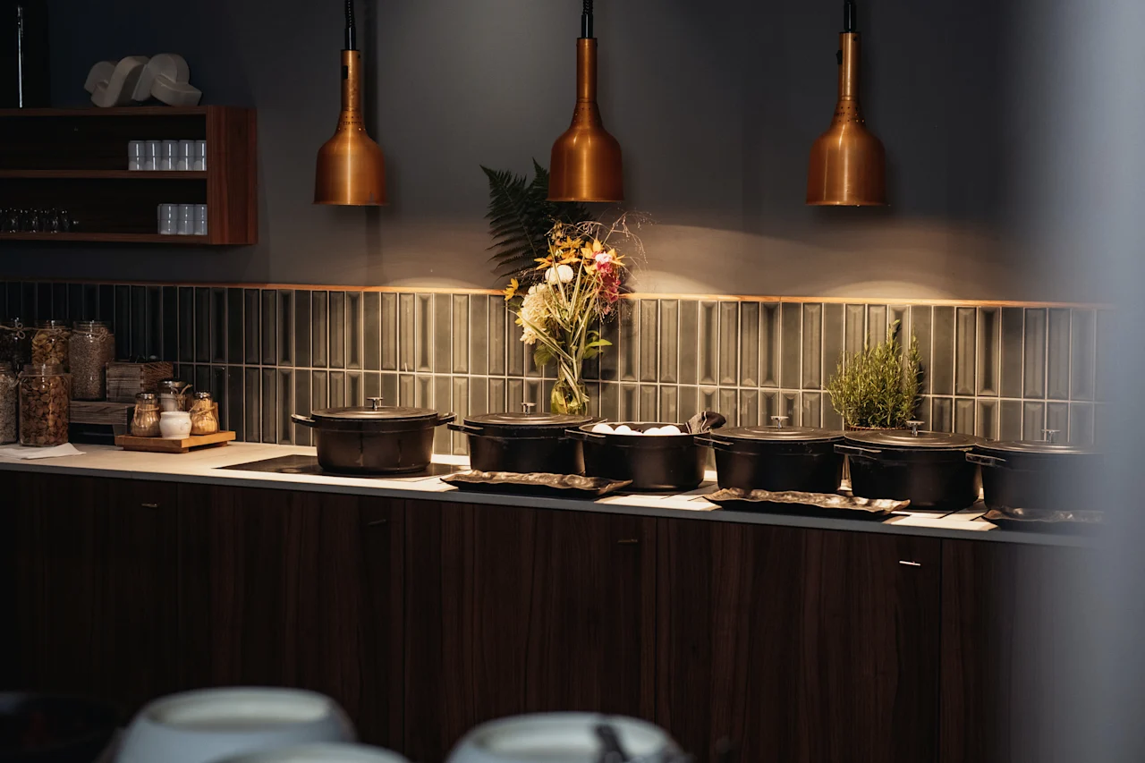 En varm och inbjudande bufféuppställning med svarta serveringsgrytor, burkar med flingor och färska blommor under kopparfärgade taklampor.