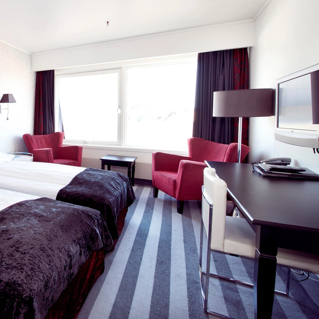 An elegantly decorated hotel room with two single beds, a red armchair, a desk, and a window.