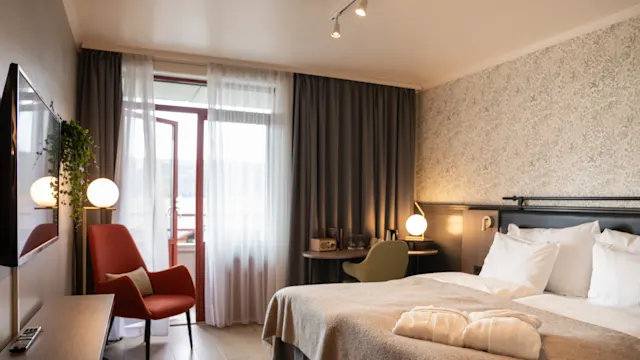 Et koselig hotellrom har en pent oppredd seng med to morgenkåper på, ved siden av en lenestol og et lite skrivebord, innrammet av store gardiner.