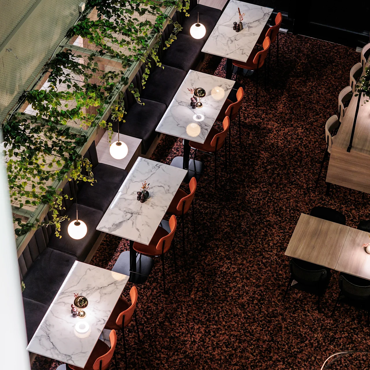 Marble-topped tables line a contemporary dining area, paired with orange chairs, surrounded by hanging greenery and warm globe lights, creating an inviting restaurant ambiance.