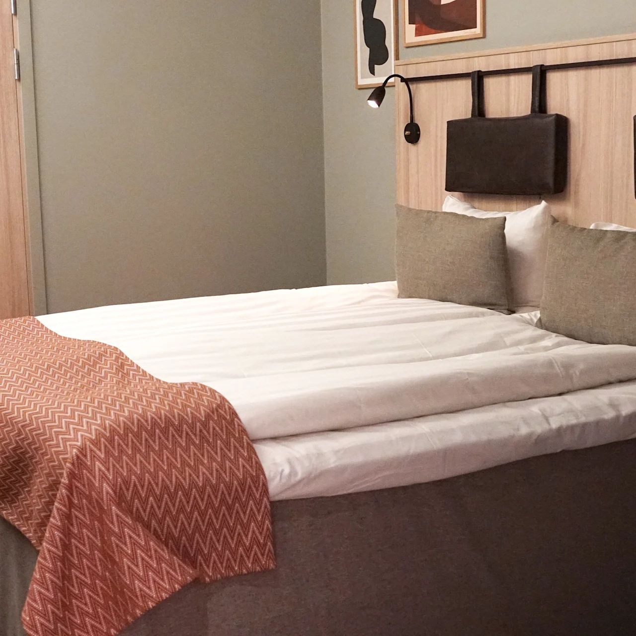 A neatly made bed with twin pillows and a rust-colored throw blanket stands against a wood-paneled wall with mounted lamps, beside a wooden door in a cozy, carpeted room.