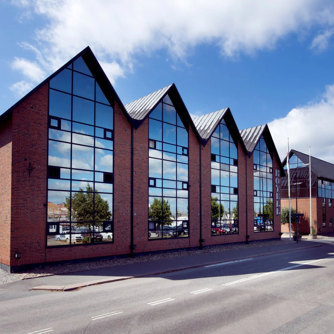 Red brick building with large windows and peaked roofs beside a street under a clear sky.