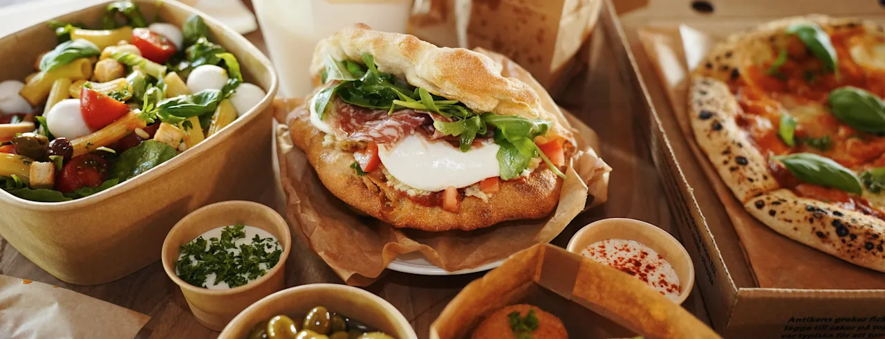 Erilaisia italialaisia ruokia, kuten pizzaa, salaattia, voileipiä ja oliiveja, on asetettu puupöydälle, mikä viittaa rentoon ruokailu- tai takeout-tilanteeseen.