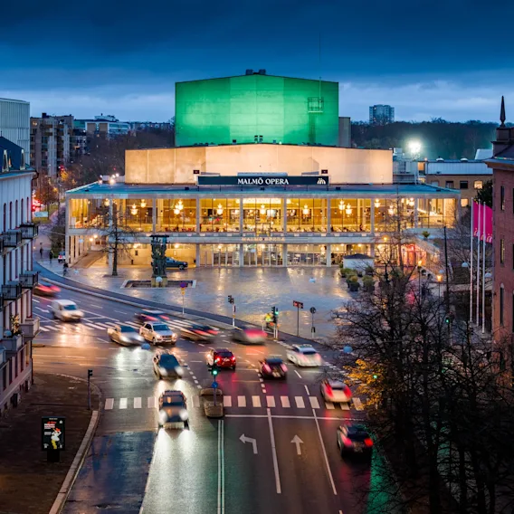 Malmo Opera upplyst i skymningen med stadstrafik som rör sig på gatan framför.