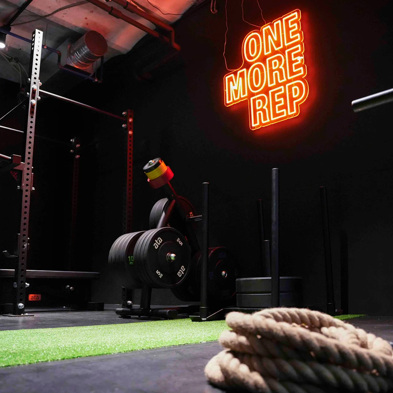 Et knebøystativ og treningsutstyr står ubrukt i et svakt opplyst treningsstudio. Neonskilt lyder "ONE MORE REP". Tykt tau ligger sammenrullet på grønt gress. Industrielt tak synlig.