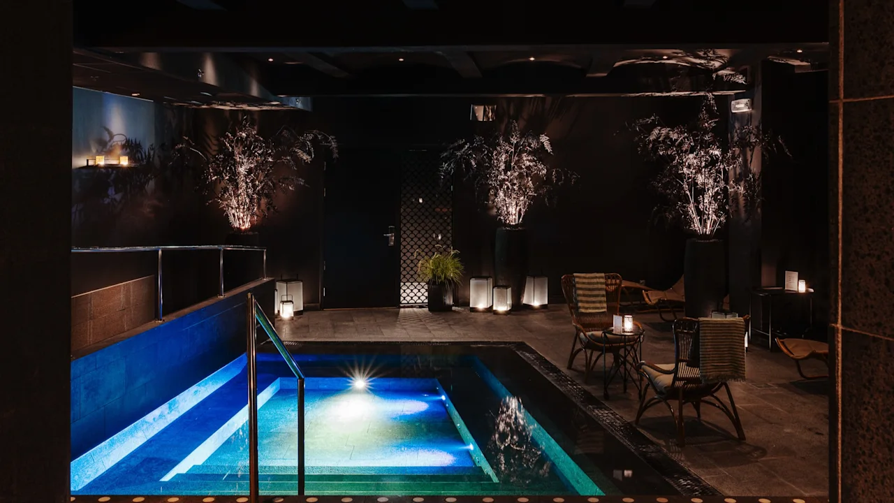 Dunkelt upplyst inomhuspool på Clarion Hotel Post med blått och grönt vatten, växter och sittplatser för avkoppling.