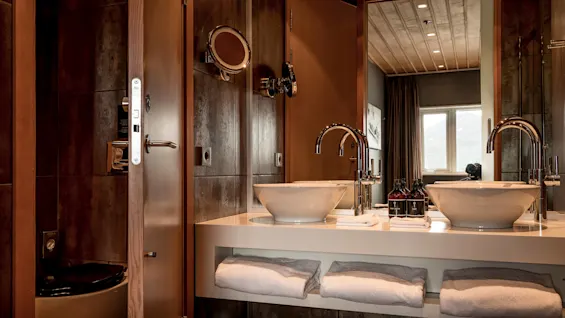 Elegant bathroom interior with a mirrored vanity, dual sinks, towels, and dark wood panels, reflecting a luxury setting.