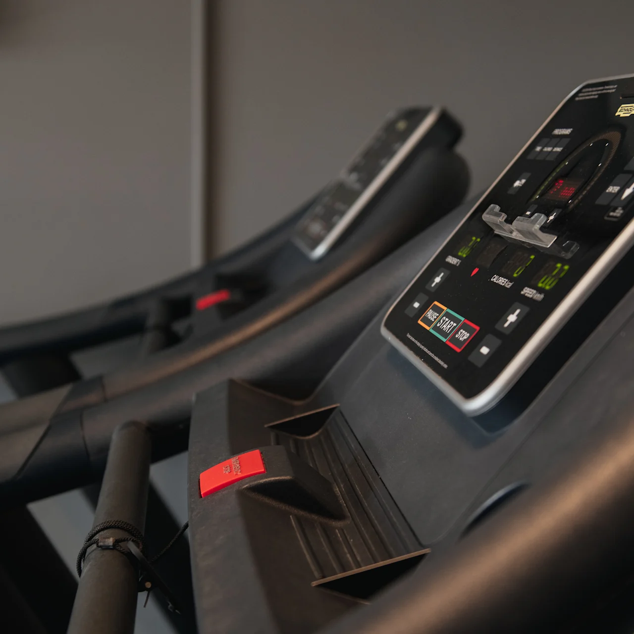 Treadmills with illuminated control panels stand stationary in a gym setting. Panels display buttons and indicators for speed and incline. Gray walls form the backdrop.