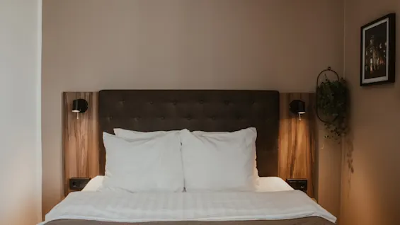 Viihtyisä hotellihuone siististi pedatulla sängyllä, valkoisilla tyynyillä ja lämpimällä valaistuksella, kutsuen rentouttavaan oleskeluun.