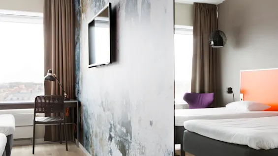 Et moderne hotelværelse har en seng, en vægmonteret TV og et lille skrivebord med stol og lampe. Store vinduer med udsigt over en by.