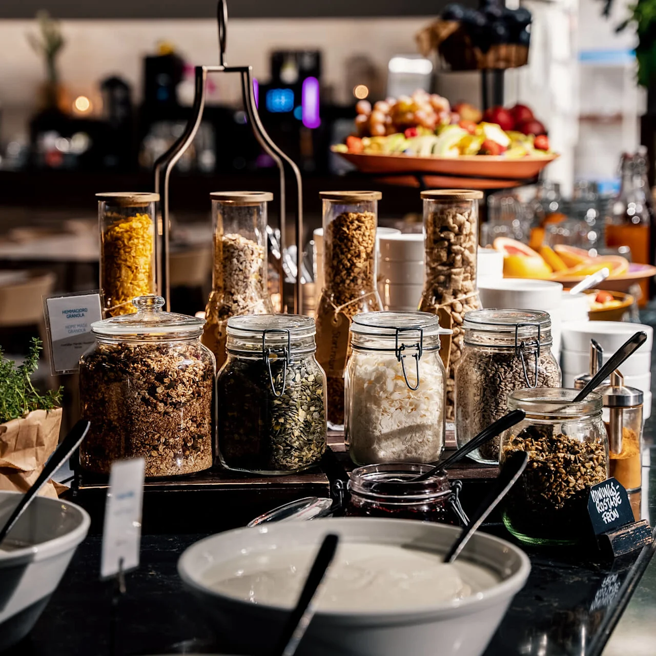 Bottles and jars filled with granola and seeds displayed on a breakfast buffet. Bowls of yogurt and fruit in the background. Labeled jar reads "Håndlavet Granola 50% Rawnola."