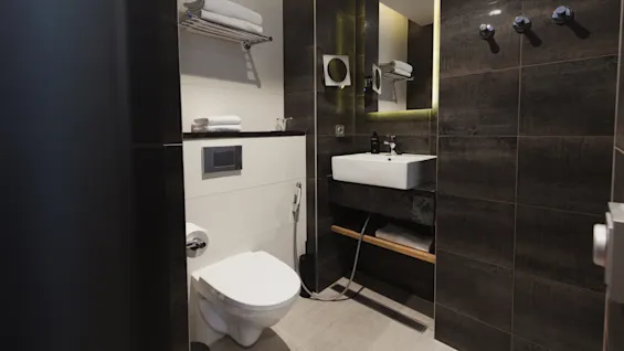 Toilet installeret på en flisevæg, placeret nær en vask under et spejl, omgivet af foldede håndklæder på hylder i en moderne badeværelsesindretning.