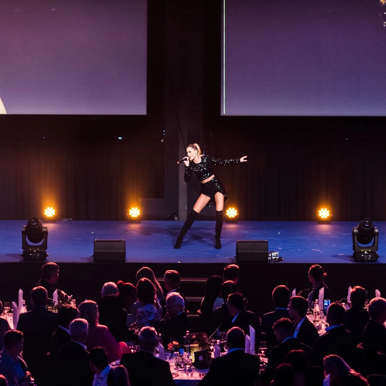 A performer in a black outfit sings energetically on stage, surrounded by bright lights, in front of a seated audience at a formal event.