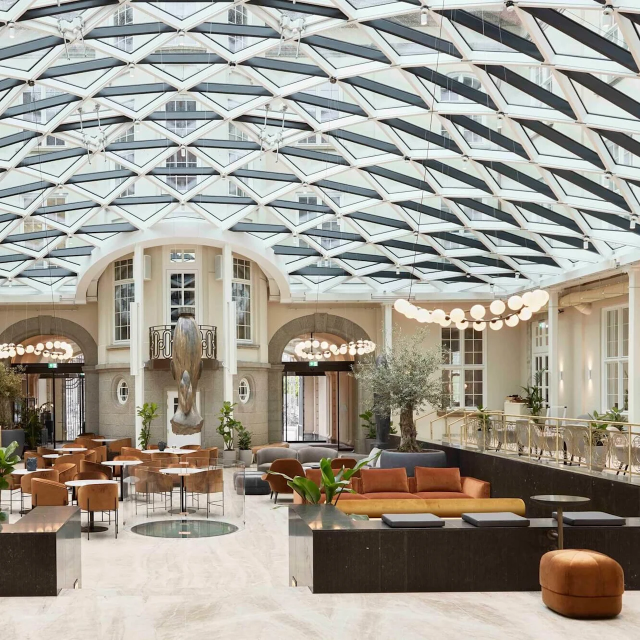 A spacious lobby features modern seating arrangements and a central statue beneath a geometric glass ceiling, surrounded by lush plants and warm lighting.