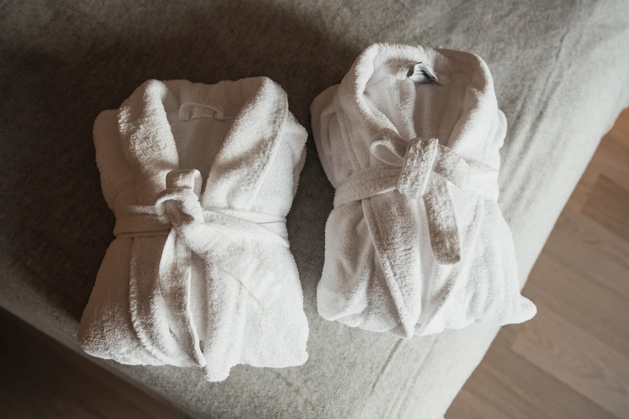 To bløde hvide badekåber pænt foldet på en seng, klar til brug, hvilket antyder komfort og afslapning.