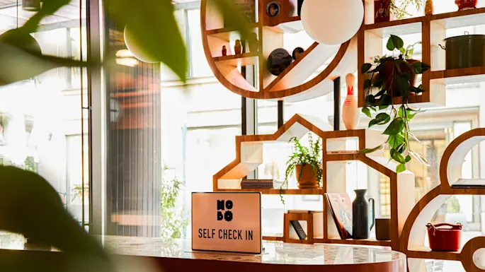 Et skilt på et bord lyder "HOBO SELF CHECK IN" i et moderne interiør med geometriske hyller, planter og dekorative gjenstander.