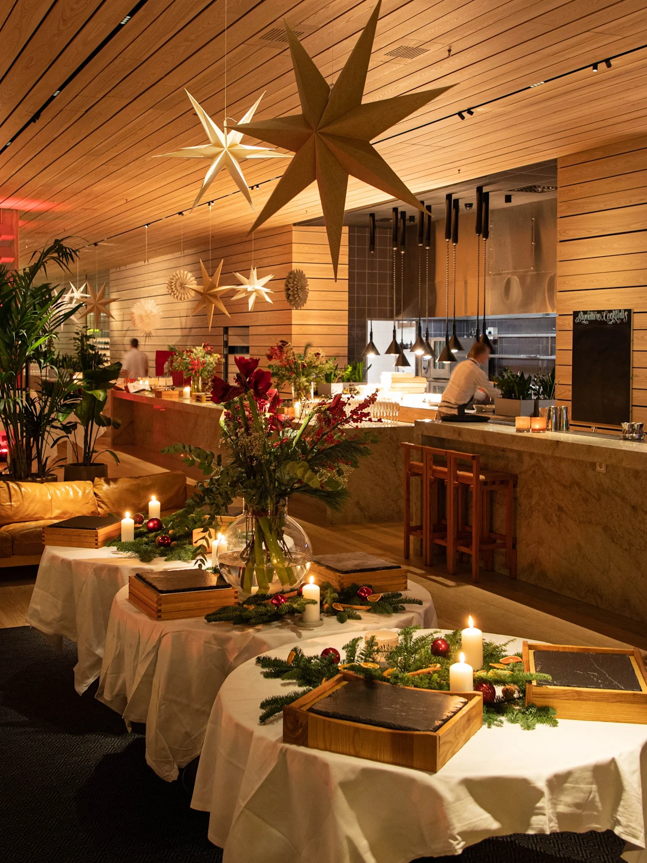 Mysig restaurang med träinredning har stjärnformade lampor över bord dekorerade festligt. En bartender serverar drycker vid en lång bardisk med flaskhyllor. Varm belysning skapar en inbjudande atmosfär.