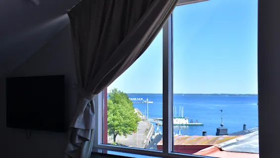 En naturskön utsikt från ett fönster över ett lugnt blått hav med en hamn, båtar och gröna träd under en klar himmel.
