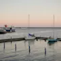 Sejlbåde fortøjet i en rolig marina under en blød solnedgang, med stille vand og en fjern kystlinje.