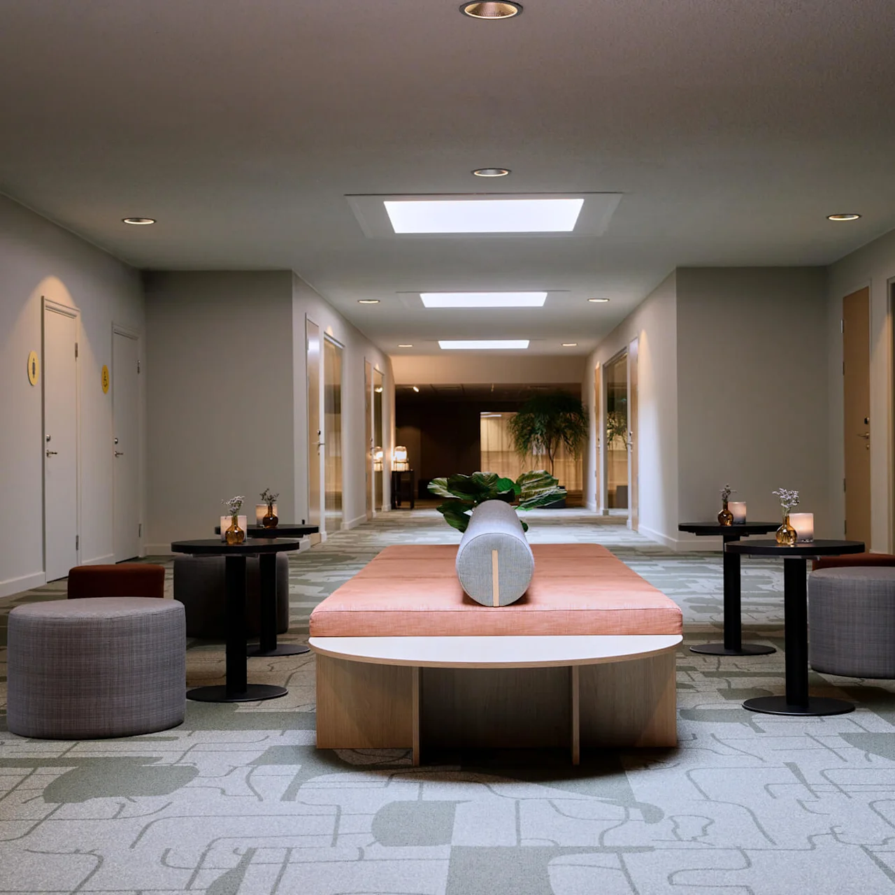 A long bench with pillows sits in a modern hallway, flanked by round stools and small tables. Potted plants and doors line the carpeted space.