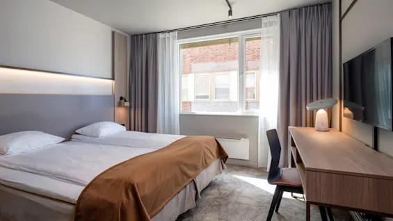 Seng med brun tæppe står ubrugt i et hotelværelse, med skrivebord, stol og vindue med grå gardiner delvist åben, og afslører en udsigt til en murstensbygning.