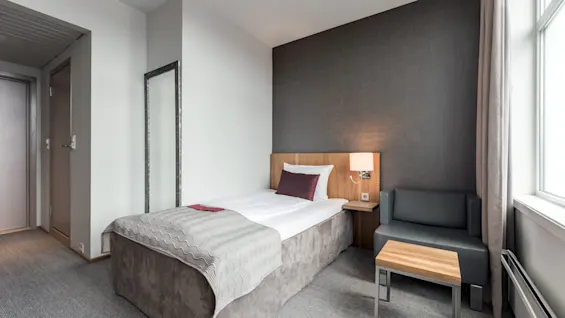 Yksi sänky, jossa on valkoiset liinavaatteet ja violetti tyyny, on harmaan sohvan ja puupöydän vieressä modernissa hotellihuoneessa, jossa on kokolattiamatto ja lattiavalaisin.