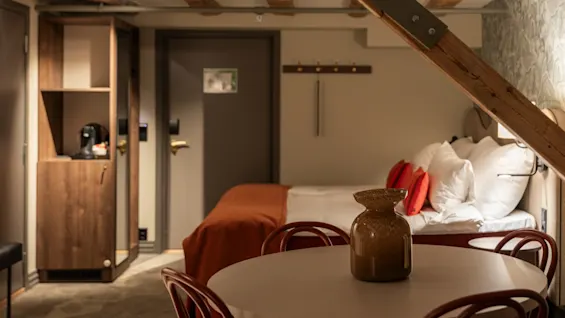 Et koselig hotellrom med seng, en liten spiseplass og synlige trebjelker, som skaper en varm og innbydende atmosfære.