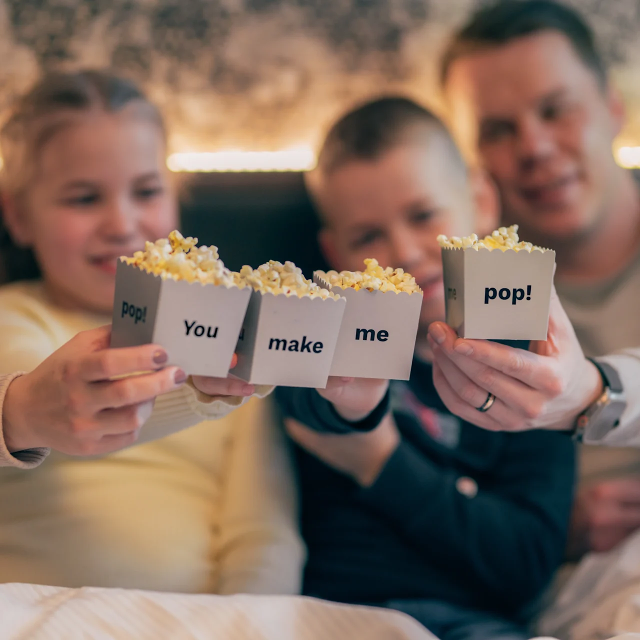 Four people hold popcorn boxes with text: "pop! You make me pop!" while sitting closely together, smiling in a cozy home setting.