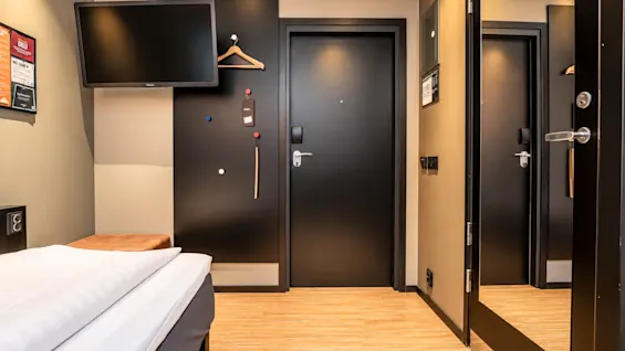 Modern hotel room entrance featuring wooden flooring, a black door, wall-mounted TV, and a large mirror.
