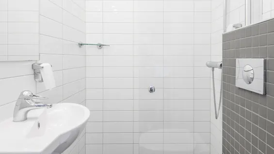 Ett ljust, modernt badrum med ett vitt handfat och en elegant kran, en väggmonterad tvålpump och en dusch. Vita kakel täcker väggarna och skapar en ren, minimalistisk atmosfär.