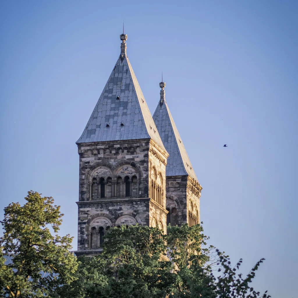 The majestic spires of Lunds domkyrka rise above green trees against a clear blue sky, bathed in sunlight.