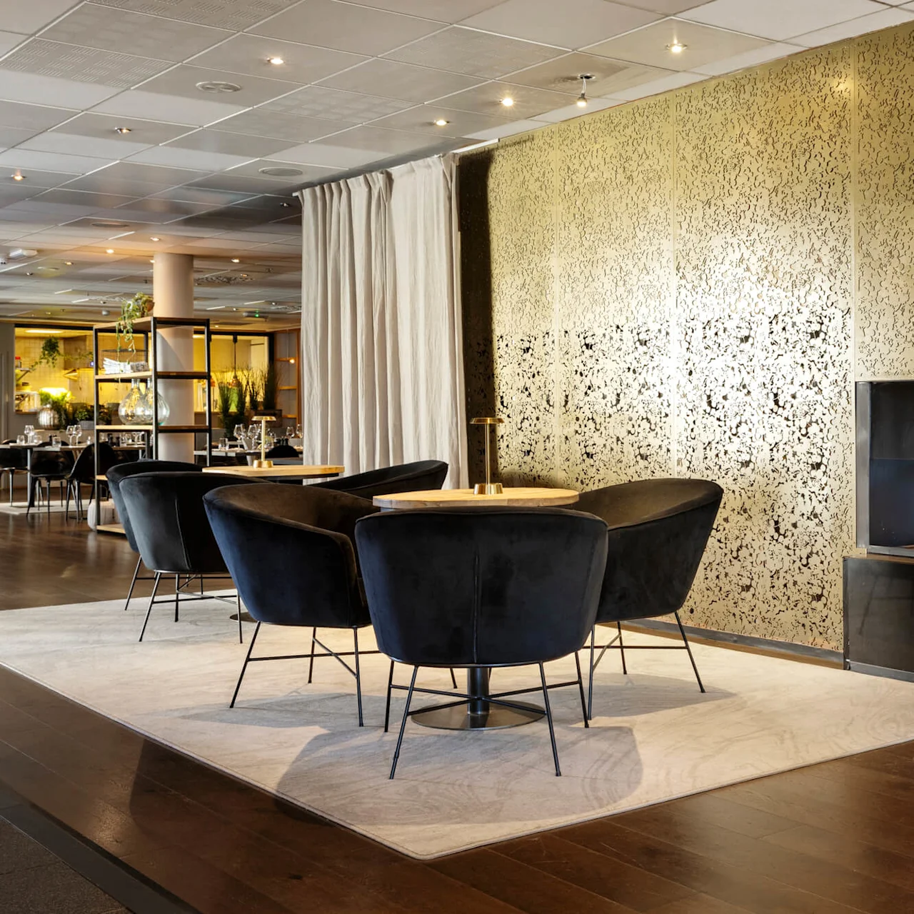 Sleek black chairs surround round tables on a light rug, near a modern fireplace in a stylish restaurant with decorative golden panels and soft lighting.