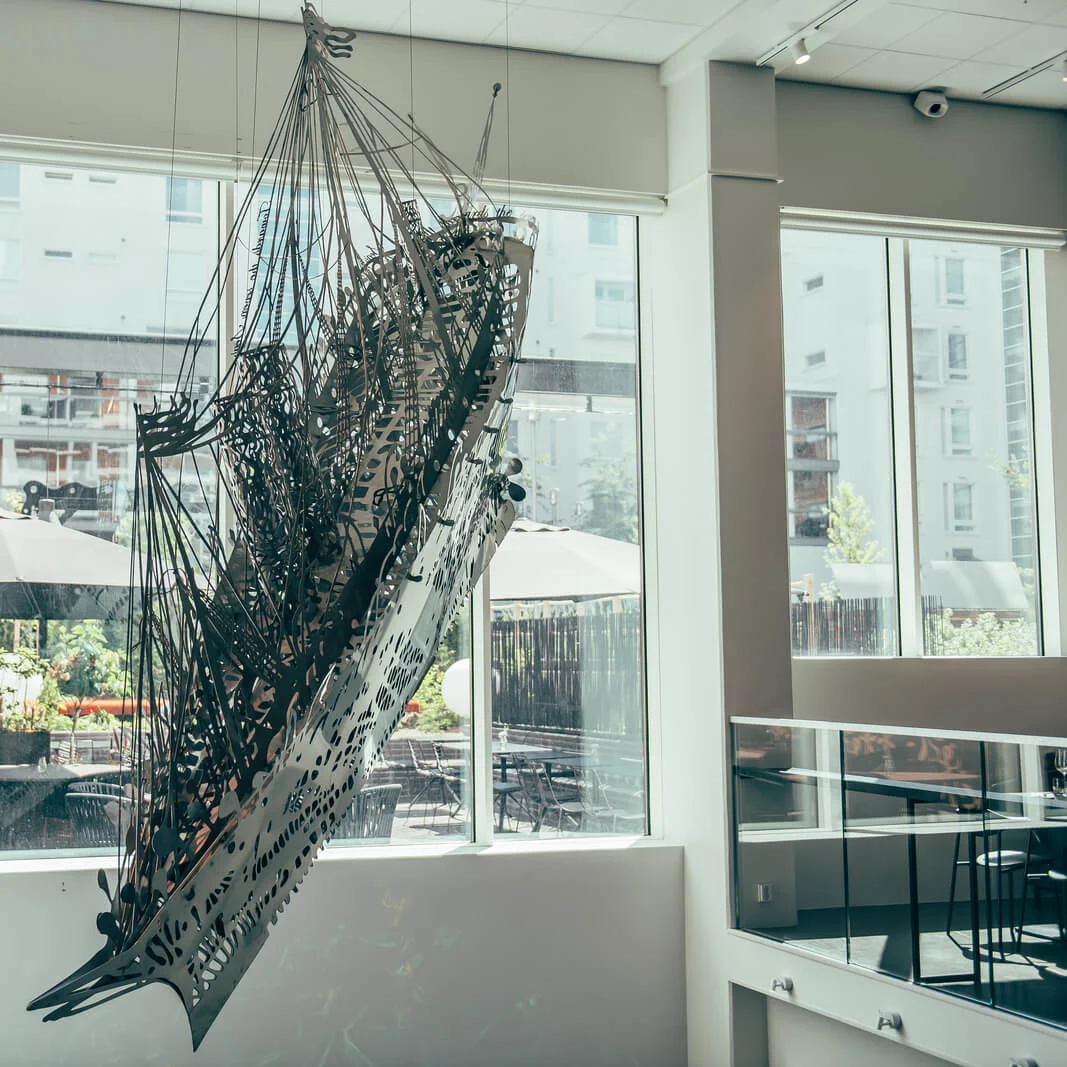 A metallic abstract sculpture is suspended in an indoor modern space with large windows overlooking an outdoor patio area with tables and umbrellas.