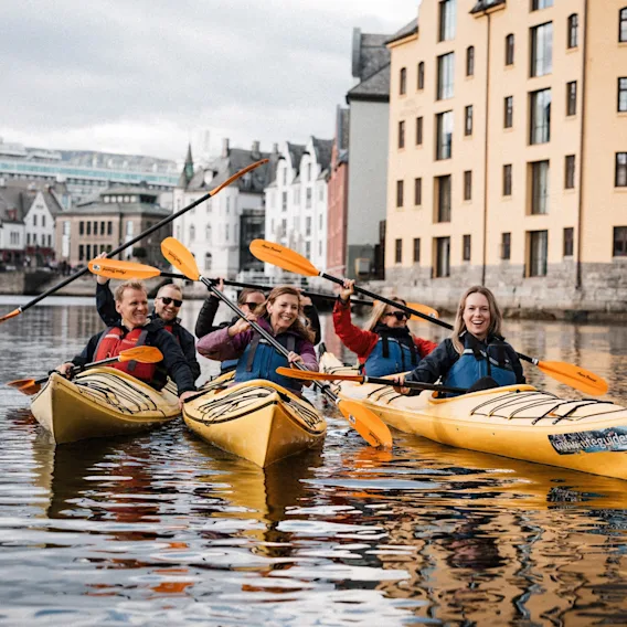 People in kayaks on a canal, smiling and enjoying the activity with buildings in the background, including a hotel.