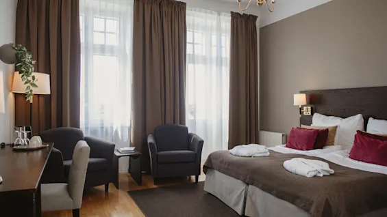 Et hotelværelse med en pænt redt dobbeltseng med hvide håndklæder, to lænestole mod et vindue og et skrivebord til siden, hvilket skaber en hyggelig og indbydende atmosfære.
