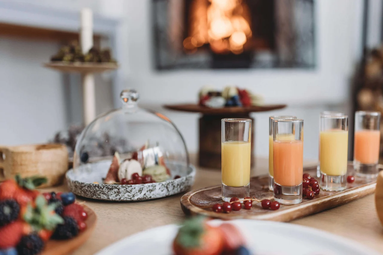 Glas med juice står på et træfad, ved siden af en kuppel-dækket tallerken med frugt og en sløret baggrund med en hyggelig pejs.