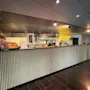 En modern bar- och receptionsdisk med snacks, drycker och skyltar för mat- och dryckesupphämtning samt delitjänster.