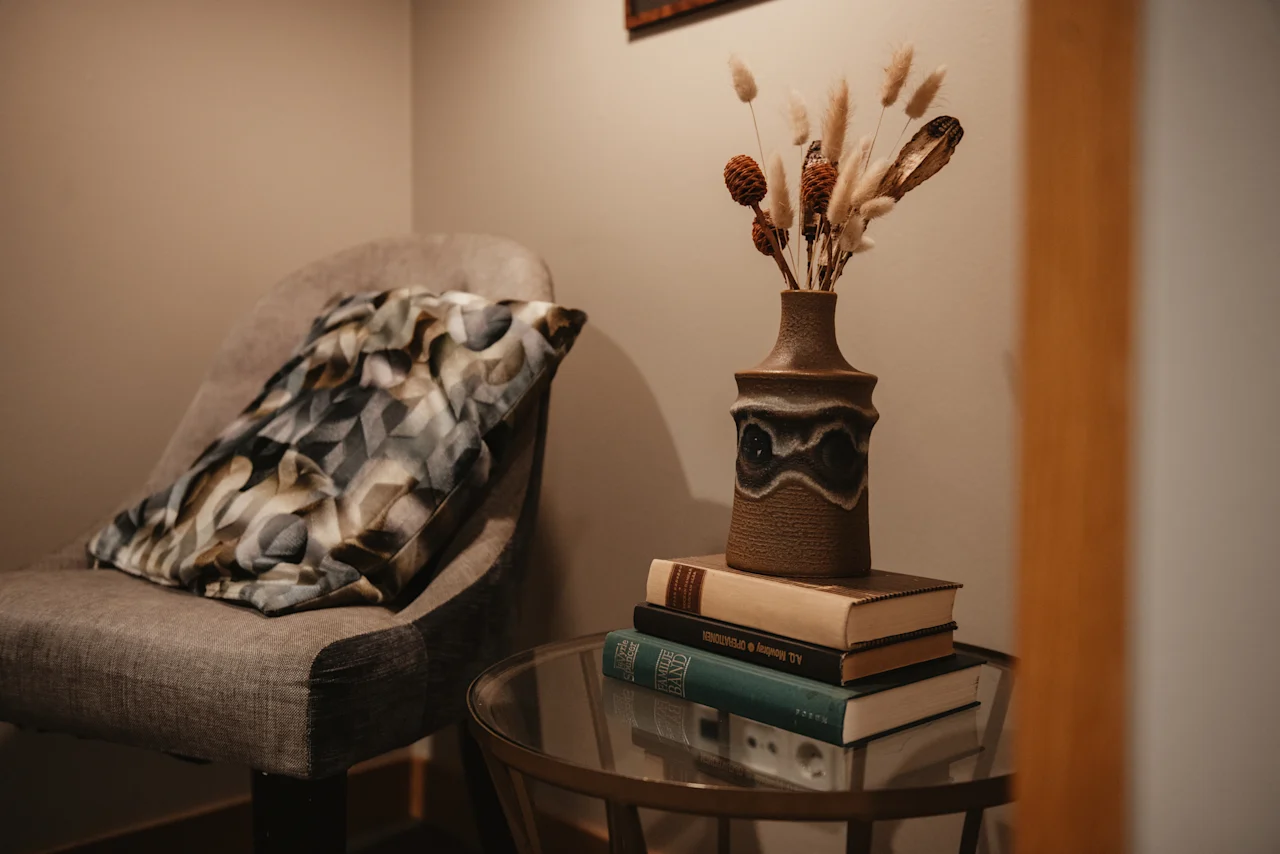 A cozy corner at Home Hotel Uppsala with a comfortable chair, decorative pillow, and a small table holding books and a vase with dried plants.