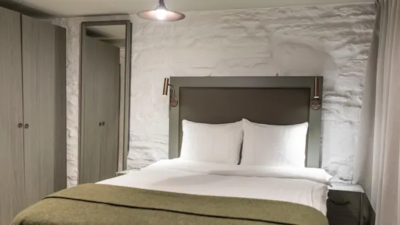 Viihtyisä hotellihuone, jossa on mukava sänky valkoisilla liinavaatteilla, vihreä peitto ja tekstuuriset kiviseinät.