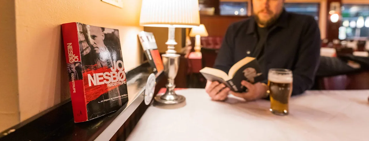 En person leser en bok ved et bord med et glass øl. En bok med tittelen RODSTRUPE står på en hylle i nærheten.