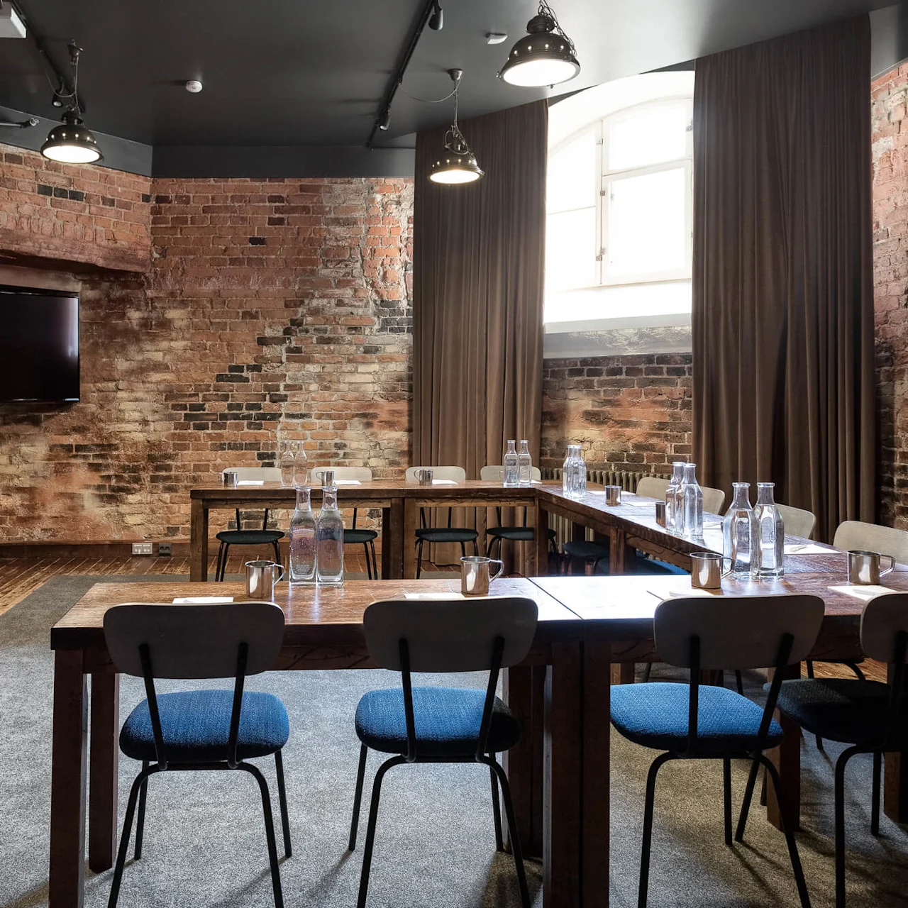 A modern meeting room with rustic elements, featuring a large wooden table, chairs, exposed brick walls, and a flat-screen TV.