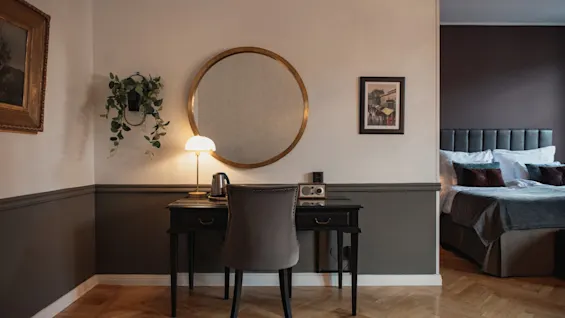 Hotellihuone, jossa on työpiste, tuoli ja suuri pyöreä peili. Sänky näkyy taustalla.