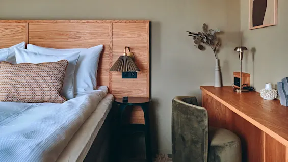 Et koselig hotellrom med en seng, en sengegavl i tre og et skrivebordsområde. En bok med tittelen 'The Art Of' ligger på et bord.