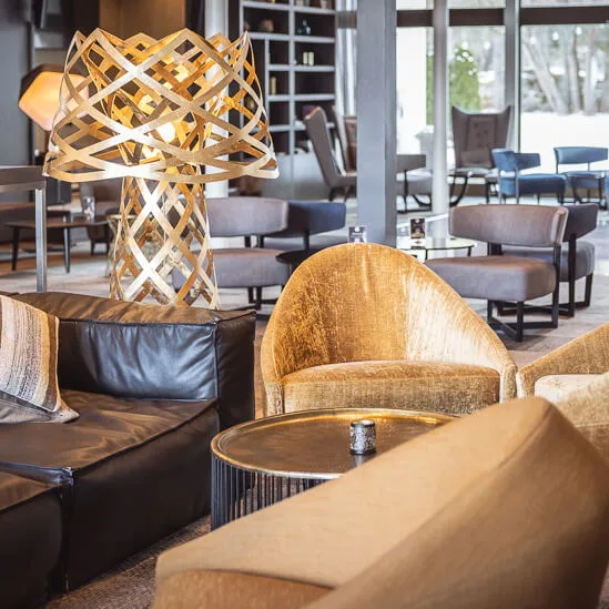 Luxurious lounge area featuring plush beige chairs surrounding a circular table. A unique, interconnected wooden lamp stands nearby. Large windows provide an outdoor view, enhancing the sophisticated atmosphere.