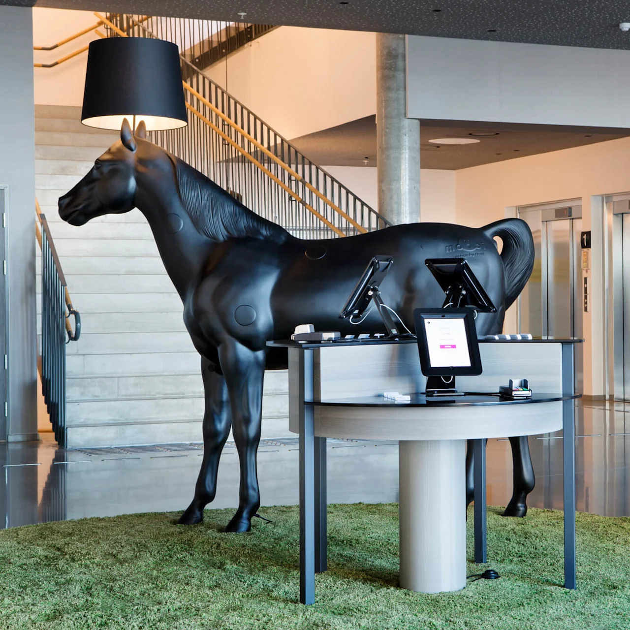 A black horse sculpture stands beneath a lamp, near stairs and elevators in a modern lobby. A circular table with electronics and green carpet accents the space.