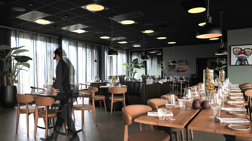 En person går gennem en moderne restaurant med dækkede borde, pendellamper og planter, hvilket antyder forberedelse til servering.
