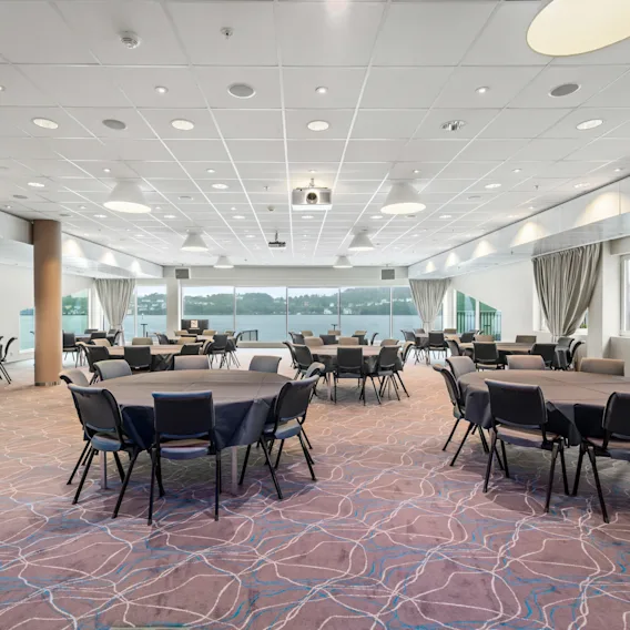 Runde bord med svarte stoler er arrangert i et romslig konferanserom. Store vinduer med gardiner slipper inn naturlig lys; et mønstret teppe dekker gulvet.